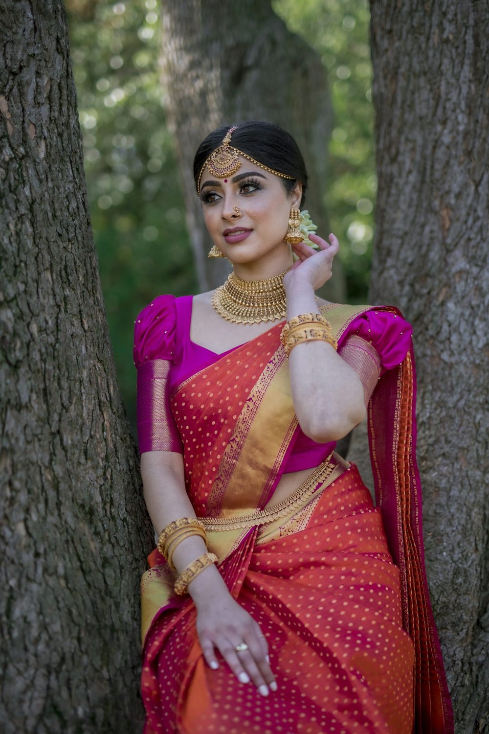A smiling woman in a pink and gold silk sari standing under the trees