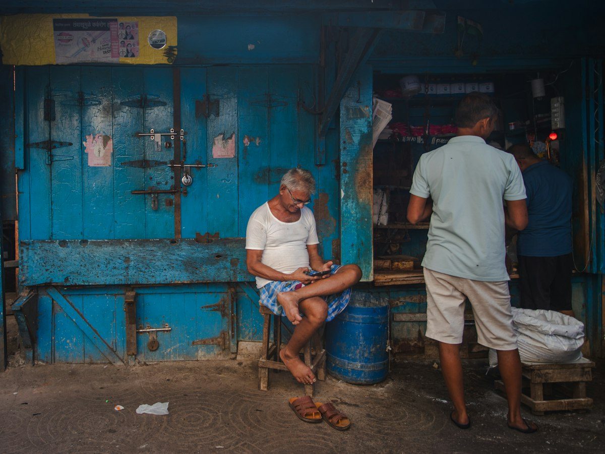 Shopkeeper sitting on a stool with his phone outside a blue shopfront